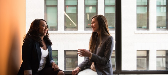Two smiling employees sit on the sill of a large office window and talk through a project in a personal and engaged way